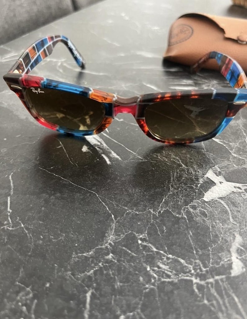 Puede incluir: Gafas de sol Ray-Ban con montura multicolor que presenta un patr&oacute;n de cuadrados azules, naranjas y marr&oacute;n oscuro. Las lentes son de color marr&oacute;n oscuro y la montura incluye detalles de metal plateado en las esquinas. Se ve un estuche de cuero beige.