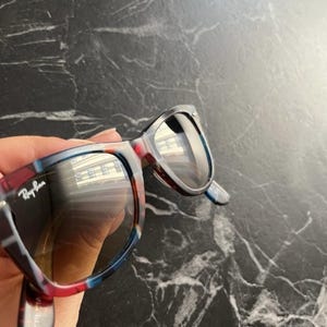 Puede incluir: Gafas de sol Ray-Ban con montura multicolor con patrones de carey en azul, rojo y marr&oacute;n. Las lentes son oscuras y reflectantes, mostrando el reflejo de un edificio. El logotipo de la marca est&aacute; impreso en blanco en la lente izquierda.