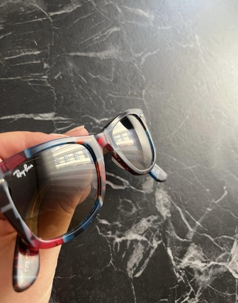 Puede incluir: Gafas de sol Ray-Ban con montura multicolor con patrones de carey en azul, rojo y marr&oacute;n. Las lentes son oscuras y reflectantes, mostrando el reflejo de un edificio. El logotipo de la marca est&aacute; impreso en blanco en la lente izquierda.