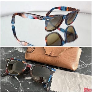 Puede incluir: Gafas de sol Ray-Ban con montura de carey multicolor en tonos azules, rojos y marrones, lentes oscuras y remaches plateados. Incluye un estuche protector de textura de cuero color canela y un pa&ntilde;o de limpieza de microfibra gris.