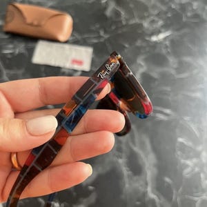 Puede incluir: Gafas de sol Ray-Ban con un patr&oacute;n de carey multicolor en la patilla y el logotipo de la marca impreso en blanco. La montura muestra una mezcla de tonos marr&oacute;n oscuro, azul y rojo. Un estuche protector de color tostado se ve al fondo.