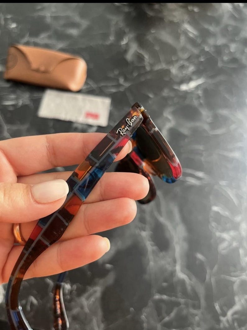 Puede incluir: Gafas de sol Ray-Ban con un patr&oacute;n de carey multicolor en la patilla y el logotipo de la marca impreso en blanco. La montura muestra una mezcla de tonos marr&oacute;n oscuro, azul y rojo. Un estuche protector de color tostado se ve al fondo.
