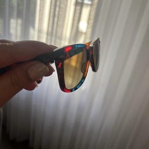 Puede incluir: Gafas de sol con montura multicolor que presenta patrones de carey en rojo, azul y marr&oacute;n. Las lentes tienen un tono amarillo dorado c&aacute;lido. El logotipo de Ray-Ban es visible en la patilla y en la esquina superior de la lente derecha.