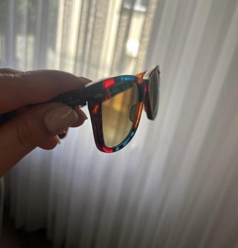 Puede incluir: Gafas de sol con montura multicolor que presenta patrones de carey en rojo, azul y marr&oacute;n. Las lentes tienen un tono amarillo dorado c&aacute;lido. El logotipo de Ray-Ban es visible en la patilla y en la esquina superior de la lente derecha.