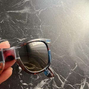 Puede incluir: Gafas de sol de carey con montura estampada en azul y marr&oacute;n oscuro. La patilla interior presenta el texto blanco Made in Italy 5 CE y 122 3N. Las lentes est&aacute;n tintadas en oscuro y la montura incluye un peque&ntilde;o detalle met&aacute;lico plateado.