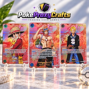 May include: Three One Piece trading cards featuring Monkey D. Luffy, Portgas D. Ace, and Sabo. Each card has a vibrant red and orange holographic background with character illustrations, game text, and power stats. The cards are displayed on clear stands.
