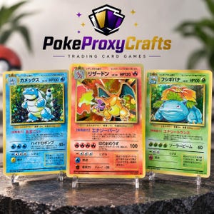 May include: Three gold-colored trading cards featuring Blastoise, Charizard, and Venusaur with Japanese text and holographic details. The cards are displayed on clear stands beneath a logo reading PokeProxyCrafts Trading Card Games.