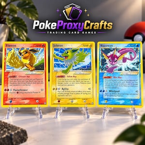 Può includere: Tre carte collezionabili con Flareon, Jolteon e Vaporeon su supporti in plastica trasparente. Ogni carta mostra un'illustrazione, 70 HP e testo su Poke-Power e attacchi. Il marchio PokeProxyCrafts è visibile nella parte superiore.