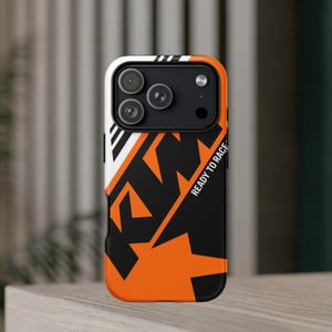 May include: A black and orange phone case featuring the KTM logo and the text READY TO RACE. The design includes bold geometric shapes and a prominent camera cutout area at the top, showcasing a modern, high-contrast aesthetic for a mobile device.
