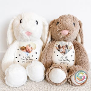Puede incluir: Dos conejos de peluche, uno blanco y otro marrón, cada uno con una impresión fotográfica en forma de corazón en el pecho. El conejo blanco muestra el texto A Cuddle From The Stars Love Grandad, y el marrón A Cuddle From The Stars Love Nanny.