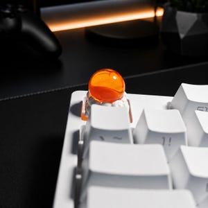 May include: A custom mechanical keyboard keycap featuring a translucent orange sphere resting on a white cloud-like base, installed on a white keyboard. The keycap is positioned among standard white keys, showcasing a smooth, glossy finish.