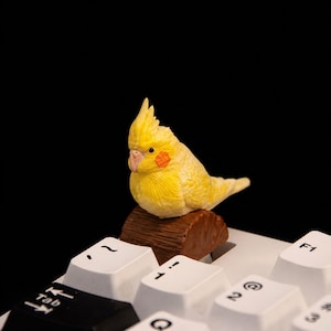May include: A yellow cockatiel figurine with orange cheek patches perched on a brown log-shaped base, positioned on top of a white computer keyboard key. The bird has a crest on its head and a light-coloured beak, resting above the Tab and 1 keys.