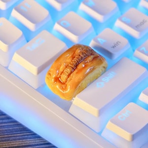 May include: A custom artisan keycap shaped like a glazed hot dog with a textured brown sausage and yellow bun, positioned on a white mechanical keyboard with blue backlit keys. The keycap replaces a standard key, featuring a glossy finish.