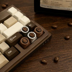 May include: A mechanical keyboard featuring a brown frame and a mix of cream, beige, and dark brown keycaps. Three custom artisan keycaps are integrated, each displaying a single roasted coffee bean embedded in a circular silver or wood-toned base.