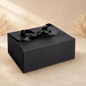 Könnte beinhalten: Eine rechteckige schwarze Geschenkbox mit glatter, matter Oberfläche und einem breiten schwarzen Satinband, das zu einer dekorativen Schleife auf dem Deckel gebunden ist. Die Box hat klare, scharfe Kanten und eine durchgehend schwarze Farbe.