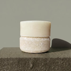 May include: Two stacked circular bars of solid soap. The top bar is smooth and off-white, while the bottom bar has a textured, granular surface with a slightly darker beige tone. Both bars are cylindrical in shape and appear to be solid cleansing products.