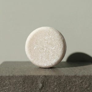 Può includere: Una saponetta solida per capelli circolare di colore bianco sporco con una superficie ruvida e granulosa. Il prodotto detergente a forma di disco presenta un aspetto compresso e opaco con piccole particelle visibili in tutta la sua composizione.