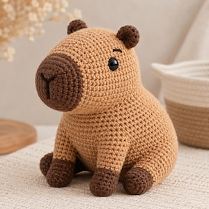 May include: A crocheted capybara plush toy featuring a light tan body, a darker brown snout, and dark brown paws and ears. The stuffed animal has small black plastic eyes and a stitched nose detail, crafted with a visible yarn texture throughout.
