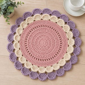 Pode incluir: Um tapete redondo de crochê com um centro rosa empoeirado e um padrão circular texturizado. É contornado por um anel de pontos festonados na cor creme, seguido por uma borda externa de pontos festonados roxos em um design decorativo.