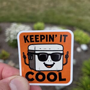 May include: A square sticker with an orange background featuring a white cooler character wearing black sunglasses. The cooler has thin black arms with fingers pointing upward. The text "KEEPIN' IT COOL" is printed in bold black letters above and below.