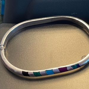 May include: A silver bangle bracelet with a multi-colored inlay of turquoise, purple, black, and green stones.