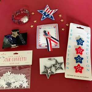 May include: Assortment of red, white, and blue star-shaped buttons and embellishments.  The buttons are labeled "Designer Button Covers" and "Connection".  The embellishments are labeled "God Bless America" and "Roblyn Designs".  There is also a package of silver star confetti labeled "New Year's Star Confetti".
