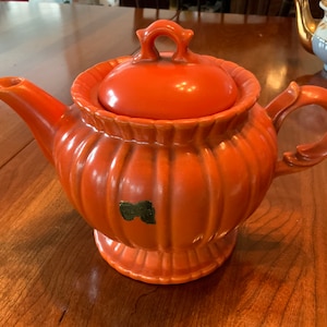May include: Orange ceramic teapot with a ribbed design and a lid. The teapot has a gold sticker on the side.