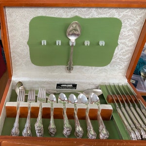 May include: A wooden box with a green velvet lining and a set of silver flatware. The flatware is arranged in a row and includes a spoon, fork, and knife. The box has a label that reads "Holmes & Edwards DeepSilver".