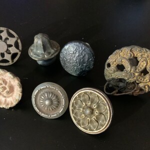 May include: Seven antique cabinet knobs with various designs. The knobs are made of metal and have a variety of finishes, including silver, gold, and black. Some of the knobs have intricate floral patterns, while others have simple geometric designs.