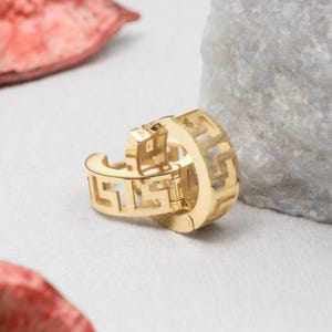 Könnte beinhalten: Ein Paar Ohrringe in Goldoptik mit einem eingravierten, sich wiederholenden geometrischen griechischen Mäandermuster auf der Außenseite. Die Ohrringe verfügen über einen Klappverschluss und zeichnen sich durch eine glänzende Oberfläche aus.