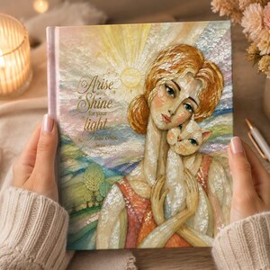 以下が含まれることがあります： 赤褐色の髪の人物が白い猫を抱いているイラストが描かれたハードカバーの本。表紙には太陽の光、丘、そして「Arise Shine for your light has come Isaiah 60:1」という文字が記されている。アートは質感のある虹色の仕上げが施されている。