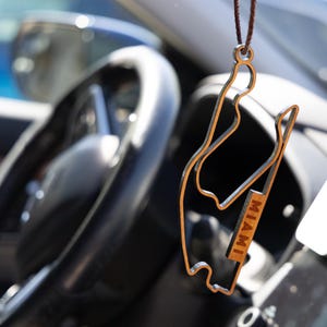 Puede incluir: Un adorno colgante de madera marrón con la forma del trazado del circuito de Fórmula 1 de Miami. El diseño recortado presenta un contorno fino del circuito con la palabra MIAMI grabada en letras mayúsculas en una sección recta del marco.