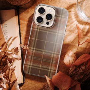 May include: A smartphone case featuring a brown, tan, and olive green plaid pattern. The case has a clear border and a square camera cutout with three circular lenses visible. The design covers the back of the phone, displaying a classic tartan-inspired grid.
