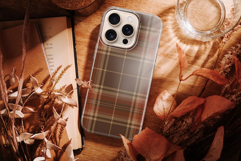 May include: A smartphone case featuring a brown, tan, and olive green plaid pattern. The case has a clear border and a square camera cutout with three circular lenses visible. The design covers the back of the phone, displaying a classic tartan-inspired grid.