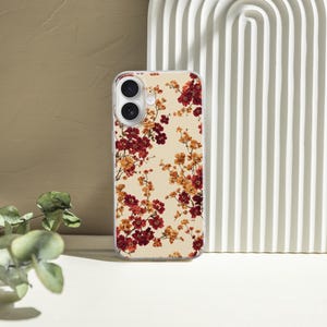 May include: A clear smartphone case featuring a repeating pattern of small red and orange flowers against a cream-colored background. The case has precise cutouts for the dual camera lenses and flash, maintaining a slim profile for the device.
