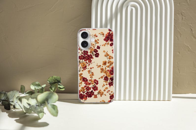 May include: A clear smartphone case featuring a repeating pattern of small red and orange flowers against a cream-colored background. The case has precise cutouts for the dual camera lenses and flash, maintaining a slim profile for the device.