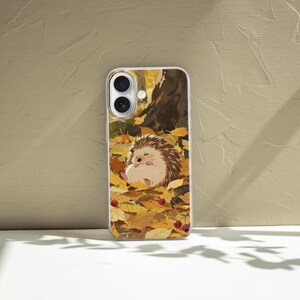 May include: A rectangular smartphone case featuring a printed illustration of a small brown hedgehog resting on a bed of fallen yellow and orange autumn leaves, with a dark tree trunk in the background and small red berries scattered throughout.