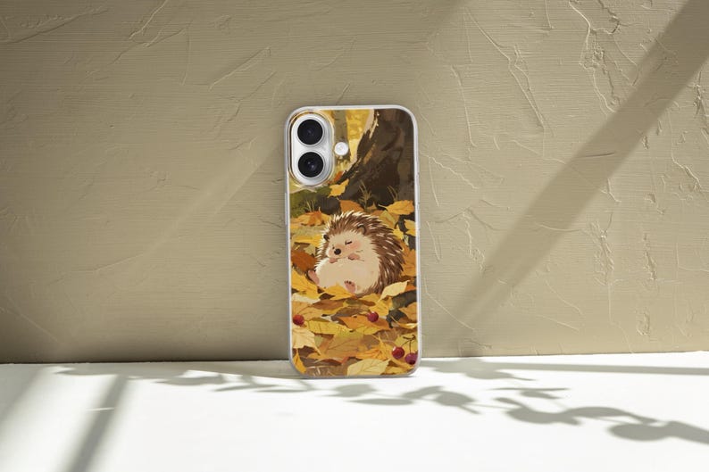 May include: A rectangular smartphone case featuring a printed illustration of a small brown hedgehog resting on a bed of fallen yellow and orange autumn leaves, with a dark tree trunk in the background and small red berries scattered throughout.