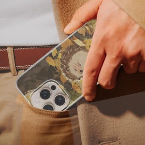 May include: A clear smartphone case featuring a printed illustration of a hedgehog surrounded by yellow and brown autumn leaves. The case is designed with a cutout for the triple-lens camera system and is shown partially inside a pocket.