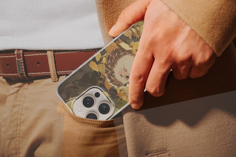 May include: A clear smartphone case featuring a printed illustration of a hedgehog surrounded by yellow and brown autumn leaves. The case is designed with a cutout for the triple-lens camera system and is shown partially inside a pocket.