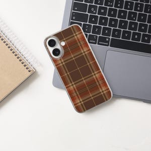 May include: A smartphone case featuring a brown, orange, and cream plaid pattern. The case has a clear border and cutouts for the dual camera lenses and flash. The design covers the entire back of the phone, showcasing a classic tartan grid.