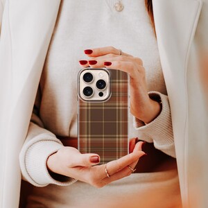 May include: A smartphone case featuring a brown, tan, and dark orange plaid pattern. The case has a clear border and a rectangular cutout for the camera lenses on the upper left side, designed to fit a device with a triple-lens camera system.