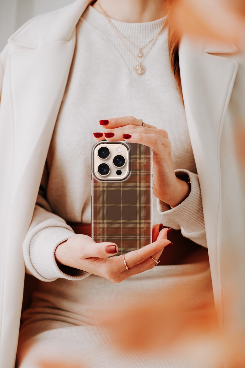 May include: A smartphone case featuring a brown, tan, and dark orange plaid pattern. The case has a clear border and a rectangular cutout for the camera lenses on the upper left side, designed to fit a device with a triple-lens camera system.