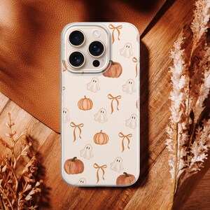 May include: A clear smartphone case featuring a repeating pattern of orange pumpkins, white ghosts, and light brown bows on a cream background. The case has a cutout for a triple-lens camera system and is designed for a modern mobile device.