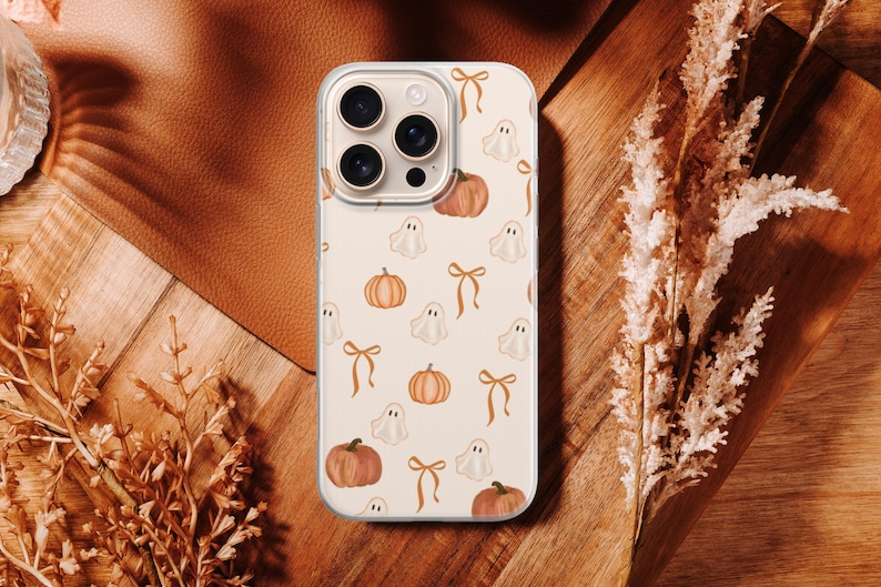 May include: A clear smartphone case featuring a repeating pattern of orange pumpkins, white ghosts, and light brown bows on a cream background. The case has a cutout for a triple-lens camera system and is designed for a modern mobile device.