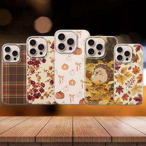 May include: Five smartphone cases featuring distinct autumn-themed designs. The patterns include a brown plaid, red and orange floral branches, white ghosts with orange pumpkins and bows, a hedgehog nestled in fallen leaves, and colourful maple leaves.