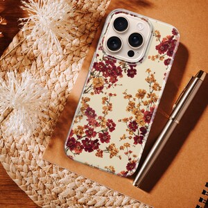 May include: A clear smartphone case featuring a floral pattern with deep red and golden yellow blossoms on a cream background. The case has a cutout for a triple-lens camera system and is positioned next to a metallic pen on a notebook.