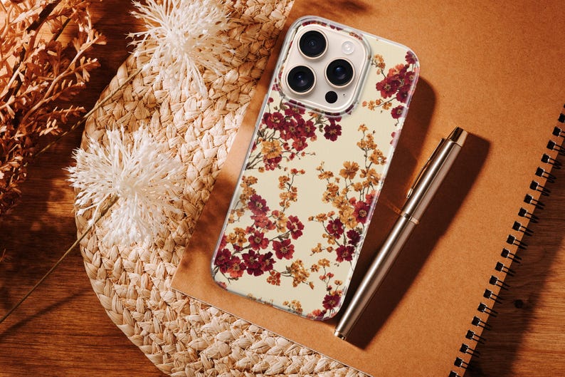 May include: A clear smartphone case featuring a floral pattern with deep red and golden yellow blossoms on a cream background. The case has a cutout for a triple-lens camera system and is positioned next to a metallic pen on a notebook.