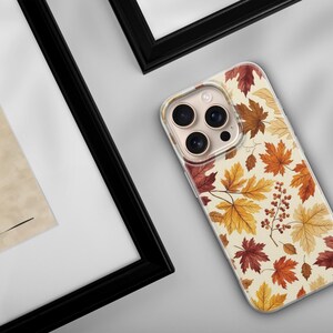 May include: A clear smartphone case featuring a repeating pattern of autumn maple leaves in shades of red, orange, and yellow, interspersed with small clusters of brown berries against a cream-colored background, shown on a device with three lenses.