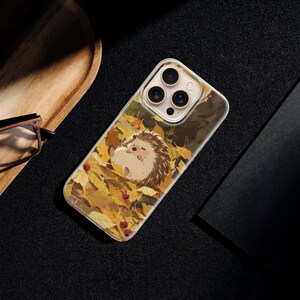 May include: A clear smartphone case featuring a graphic illustration of a small hedgehog sleeping amidst a pile of vibrant yellow and orange autumn leaves, with small red berries scattered throughout the foliage on the back of the device.